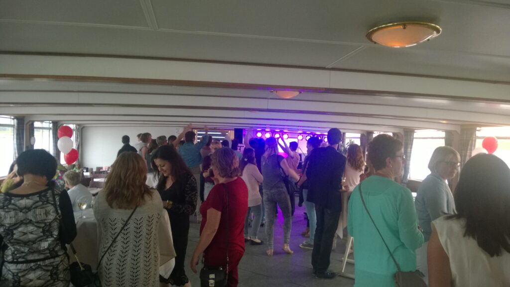 Gezellige groep mensen feestend in de binnenruimte van een partyschip, met verlichting en muziek op de achtergrond.