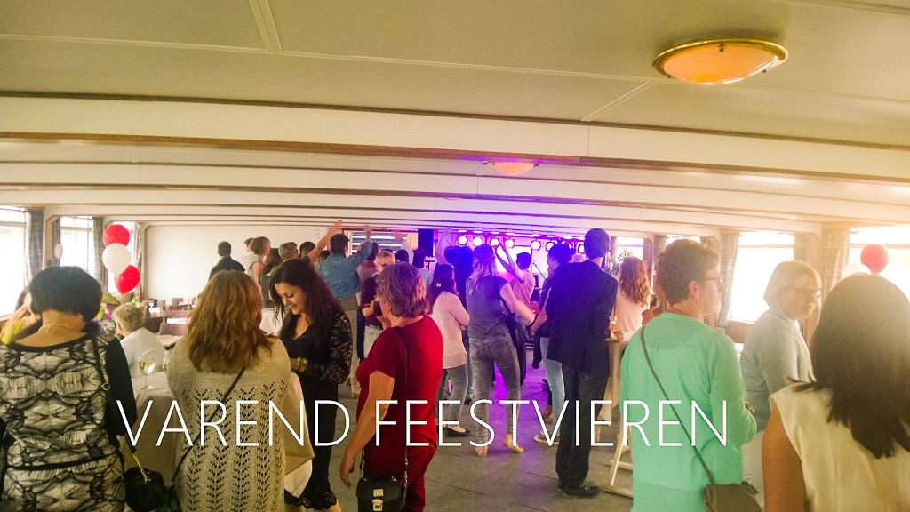 Binnenruimte van een feestschip waar gasten staan te praten en dansen, met feestverlichting op de achtergrond en de tekst “Varend Feestvieren” op de voorgrond.