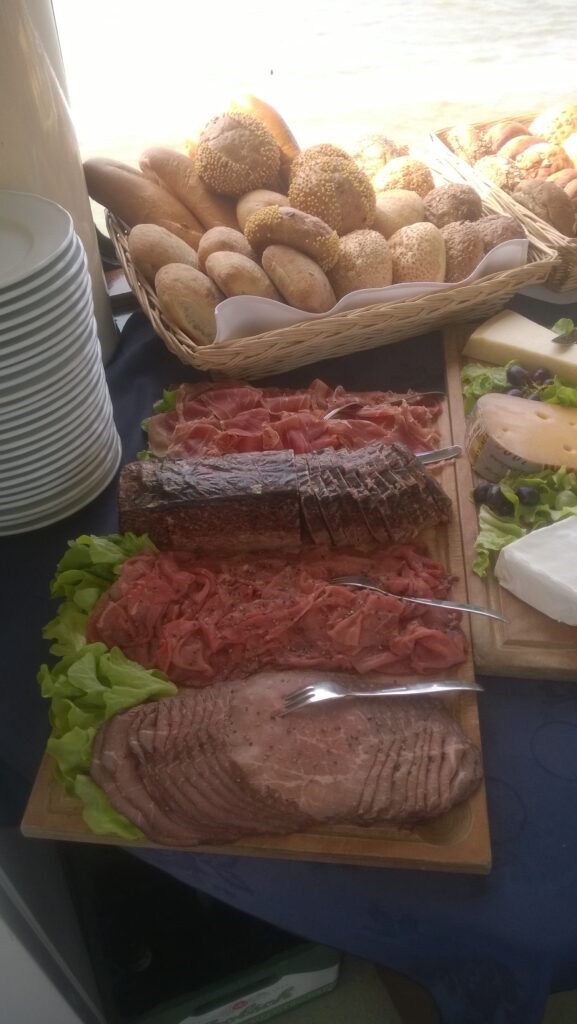 Buffettafel met een mand vol broodjes schalen met verschillende soorten vleeswaren zoals roastbeef ham en opgediend vlees omlijst met sla en naast een stapel borden