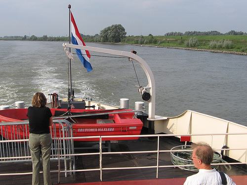 Twee personen staan op het achterdek van een passagiersschip met uitzicht op de rivier op het dek staat een rode reddingsboot en een Nederlandse vlag wappert aan de mast