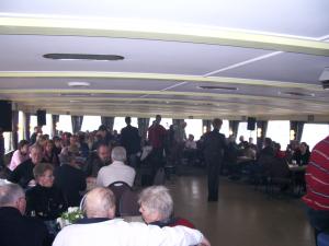 Grote groep mensen zit en staat in een drukke, overdekte scheepssalon tijdens een bijeenkomst of evenement.