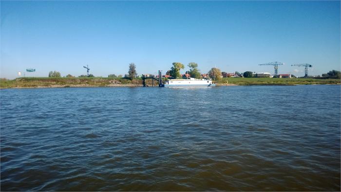 Rivierlandschap met een rondvaartboot aangemeerd aan een kleine steiger gezien vanaf het water met groene oevers bomen en enkele industriekranen op de achtergrond onder een heldere blauwe lucht